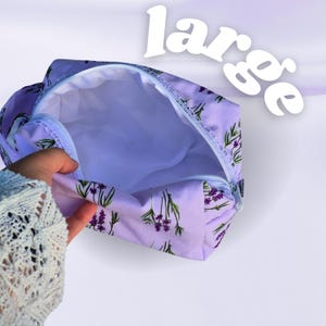 Könnte beinhalten: Ein lavendelfarbener Reißverschlussbeutel mit einem floralen Muster aus grünen Stielen und lila Blüten. Das Wort "large" ist weiß oben aufgedruckt. Der Innenraum ist ein hellerer Farbton von Lavendel.