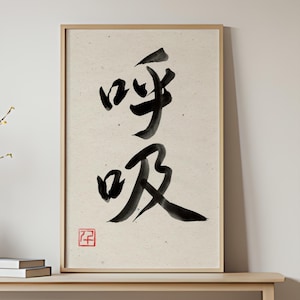 Könnte beinhalten: Gerahmter Druck mit japanischer Kalligraphie in schwarzer Tinte auf cremefarbenem Hintergrund. Das Kunstwerk zeigt zwei Schriftzeichen und ein kleines rotes Siegel. Der Rahmen ist hellbraun. Das Bild steht auf einer Holzoberfläche.