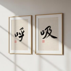 以下が含まれることがあります： 日本の書道が施された2つの額入りプリント。黒インクの文字はベージュの背景にあり、下部に小さな赤い正方形の印鑑があります。フレームは明るい木の色です。プリントは白い壁に飾られています。