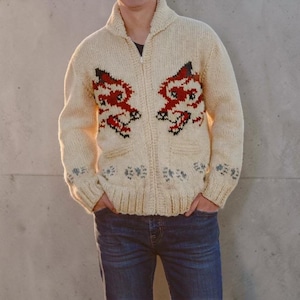 May include: Cream-coloured zip-up sweater with red and black embroidered dragon-like figures on the front. The sweater has a shawl collar, two pockets, and blue floral accents on the cuffs. The person is wearing blue jeans.