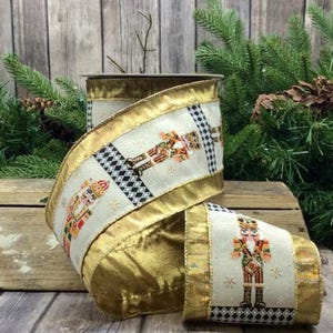 Op de afbeelding: Gouden en crèmekleurig kerstlint met een herhalend patroon van notenkrakersoldaten. Het lint heeft een metallic gouden rand en een crèmekleurig midden met het notenkrakerontwerp. Gepresenteerd op een houten oppervlak met dennentakken.