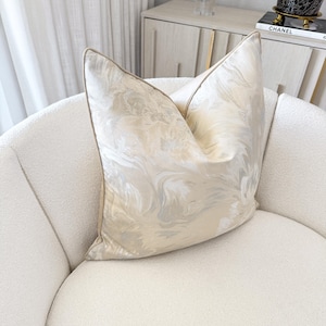 May include: A decorative pillow with a cream-colored, swirling pattern and a light brown border rests on a white, curved armchair. The pillow's design features a subtle floral motif, adding a touch of elegance to the room's decor.