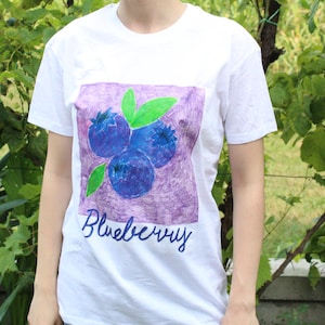 May include: A white short-sleeved t-shirt featuring a square graphic print of three blue blueberries with bright green leaves on a purple background. The word Blueberry is printed in a dark blue cursive script directly below the square graphic.