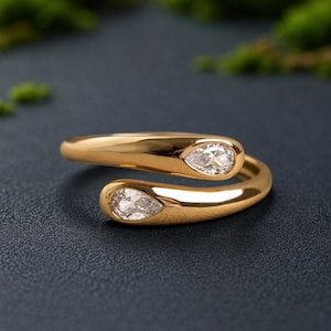 Puede incluir: Un anillo abierto de oro con dos extremos, cada uno engastado con una piedra preciosa transparente en forma de pera. La banda de oro pulido se curva para crear una silueta abierta, destacando las dos piedras talladas en forma de gota.