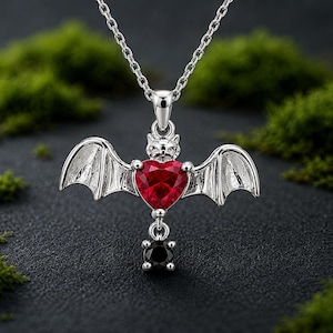 May include: A silver-toned necklace featuring a bat-shaped pendant with a faceted red heart-shaped gemstone at its center and a small, round black gemstone dangling from the bottom, suspended from a delicate silver-toned metal chain.