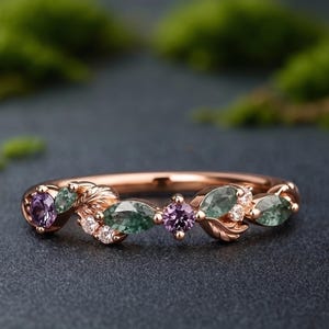 May include: Rose gold ring featuring a leaf and gemstone design. The ring has alternating purple and green gemstones, with small white accent stones. The ring is set against a dark background.