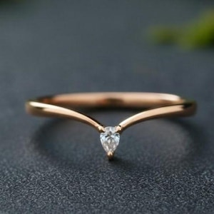 May include: A delicate rose gold ring with a V-shaped band and a pear-shaped diamond. The ring is set against a dark grey surface with blurred green foliage in the background. The ring is a piece of jewellery.