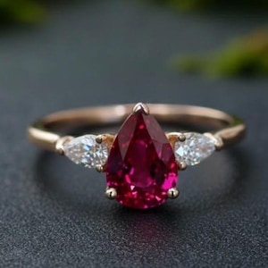 May include: A rose gold ring with a large, pear-cut, deep red gemstone at its centre. Two smaller, pear-cut, clear gemstones are either side of the main stone. The ring is set against a dark background.