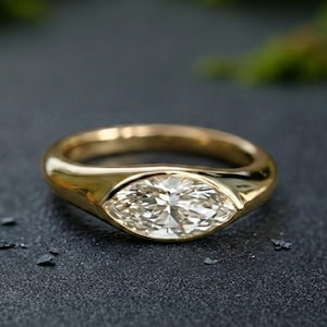 May include: A gold ring featuring a marquise-cut diamond in a bezel setting. The ring has a polished band and a sparkling diamond. The ring is set against a dark background with blurred green foliage.