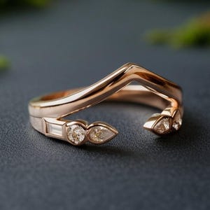 May include: Rose gold ring with a V-shaped design. The ring features three small, clear gemstones on each side. One side has a baguette-cut stone and two round stones, while the other side has three pear-shaped stones. The ring is set against a dark background.