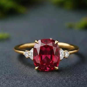 May include: A gold ring featuring a large, oval-cut ruby gemstone as the centerpiece. The ruby is a deep red color and is flanked by smaller, clear gemstones on each side. The ring is set against a dark background.