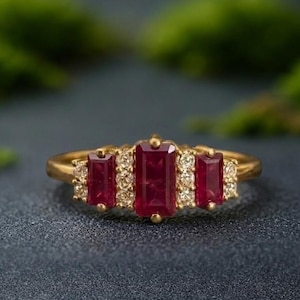 Peut inclure: Une bague en or ornée de trois pierres précieuses rectangulaires rouge foncé. La pierre centrale est plus grande que les deux pierres latérales. De petites pierres claires sont serties autour des pierres rouges. La bague est sur fond sombre.