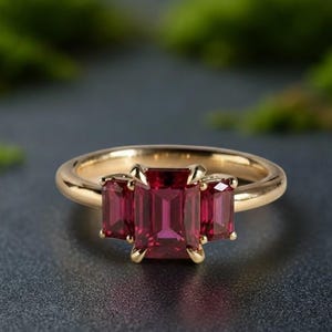 Peut inclure: Une bague en or ornée de trois pierres précieuses rectangulaires rouge foncé. La bague a un anneau en or poli et les pierres sont serties dans un design classique à trois pierres. La bague est présentée sur une surface gris foncé avec un arrière-plan vert flou.