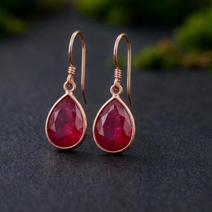 May include: A pair of drop earrings featuring faceted, teardrop-shaped deep red gemstones set in rose-colored metal frames. Each earring hangs from a simple, curved hook wire, showcasing a polished finish and a classic, symmetrical design.