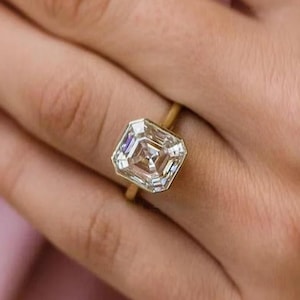 May include: A gold ring with a large, square-cut diamond. The diamond is clear and multifaceted, reflecting light. The ring's band is a thin, simple gold band. The ring is on a person's finger.