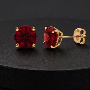 Pode incluir: Um par de brincos de tarracha com pedras preciosas redondas de cor vermelho-escura facetada em engastes de ouro amarelo de quatro garras. Um brinco é mostrado de frente, enquanto o outro exibe o perfil lateral e o fecho de pino em ouro.