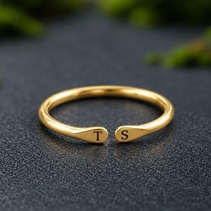 Puede incluir: Un anillo abierto de tono dorado con una banda lisa y pulida que termina en dos extremos aplanados. Cada extremo está grabado con una letra mayúscula, uno muestra la letra T y el otro la letra S, posicionadas una frente a la otra.
