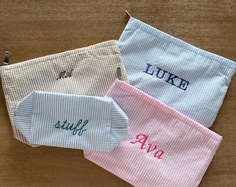 May include: Four seersucker zippered pouches in various colors. One tan pouch embroidered with MA, one light blue pouch with LUKE, one pink pouch with Ava, and one small light blue striped pouch with the word stuff embroidered in dark teal.