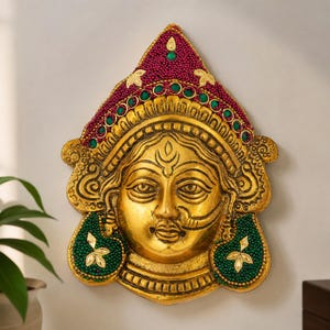 May include: A gold-toned wall plaque featuring a stylized face with a pointed crown. The crown is decorated with deep red beaded textures and small green circular accents. Two green beaded sections flank the chin, each featuring a gold leaf motif.