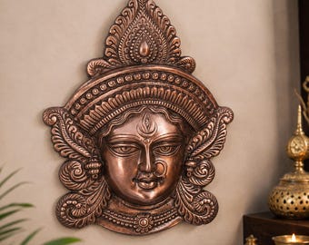 Peut inclure: Une plaque murale couleur cuivre représentant le visage sculpté d'une divinité avec un troisième œil proéminent sur le front, un anneau nasal et une coiffe ornée et superposée aux motifs tourbillonnants et aux bordures décoratives métalliques.