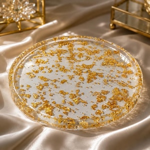 May include: A circular, transparent resin tray featuring embedded gold leaf flakes throughout the surface. The tray has a smooth, clear finish with a raised edge, showcasing the metallic gold fragments suspended within the clear material.