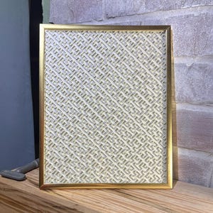 May include: A rectangular gold-framed picture with a woven, cream-colored pattern. The frame has a simple, elegant design. The picture is displayed on a wooden surface, with a brick wall in the background. The overall aesthetic is minimalist and decorative.