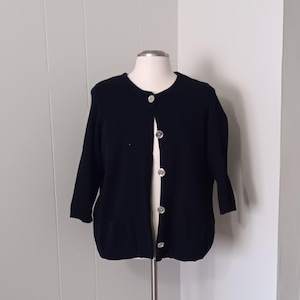 May include: A navy blue cardigan with three-quarter sleeves and a round neckline. The cardigan has large, silver-colored buttons down the front and two front pockets. The garment is displayed on a mannequin.