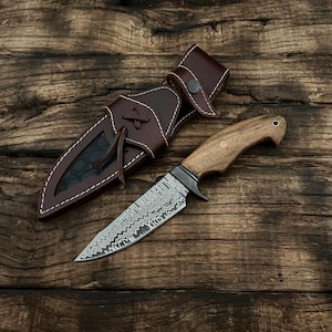 May include: A handcrafted hunting knife with a patterned steel blade and a wooden handle. The knife is accompanied by a brown leather sheath with white stitching and a snap closure. The knife and sheath are displayed on a rustic wooden surface.