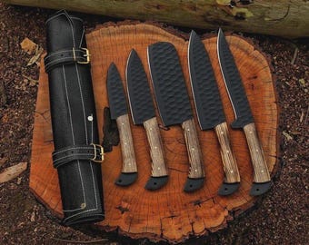 May include: A set of six black kitchen knives with wooden handles and a black leather knife roll. The knives are arranged on a wooden surface, showcasing their various blade shapes and sizes. The knife roll is closed with two buckled straps.