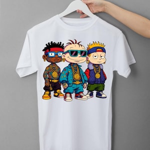Puede incluir: Camiseta blanca con un gráfico de dibujos animados de tres personajes con atuendo hip-hop. Los personajes llevan gafas de sol, cadenas de oro y trajes coloridos. La camiseta está en una percha blanca.