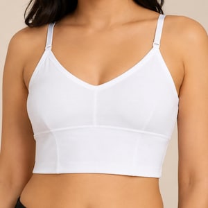Op de afbeelding: Een effen witte bralette met een V-hals, een brede band onder de buste en dunne verstelbare schouderbandjes. Het kledingstuk is voorzien van verticale naaddetails langs de voorpanelen, wat zorgt voor een gestructureerde pasvorm en een strak uiterlijk.