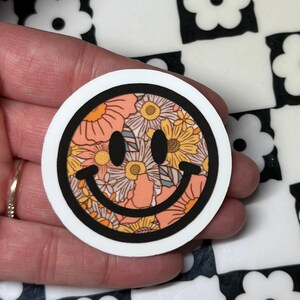 Op de afbeelding: Een ronde sticker met een smiley-gezicht ontwerp. De smiley is gevuld met een bloemenpatroon in perzik, geel en wit, omlijnd in zwart en geplaatst tegen een witte achtergrond.