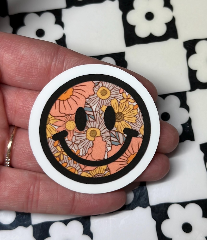 Op de afbeelding: Een ronde sticker met een smiley-gezicht ontwerp. De smiley is gevuld met een bloemenpatroon in perzik, geel en wit, omlijnd in zwart en geplaatst tegen een witte achtergrond.