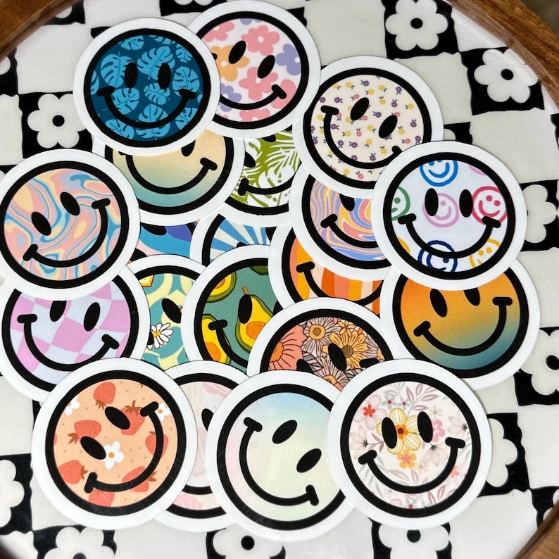 Op de afbeelding: Een verzameling ronde stickers met smiley-gezichten met verschillende patronen en kleuren. Ontwerpen omvatten bloemen, tropische bladeren en psychedelische wervelingen. Elke sticker heeft een zwarte omtrek en een witte rand, wat een vrolijke uitstraling cre&euml;ert.