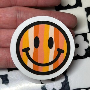 Op de afbeelding: Een ronde sticker met een lachend gezicht met een retro kleurenschema. Het ontwerp bevat verticale strepen in tinten oranje, geel en perzik, omlijnd in zwart, met een witte rand. Het smileygezicht heeft zwarte ogen en een gebogen glimlach.