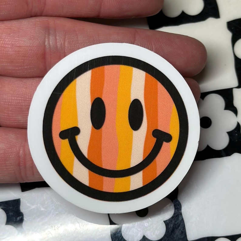 Op de afbeelding: Een ronde sticker met een lachend gezicht met een retro kleurenschema. Het ontwerp bevat verticale strepen in tinten oranje, geel en perzik, omlijnd in zwart, met een witte rand. Het smileygezicht heeft zwarte ogen en een gebogen glimlach.