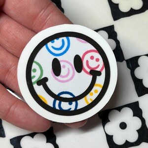 Op de afbeelding: Een ronde witte sticker met een zwarte rand met een lachend gezicht. Het gezicht is gevuld met kleinere, kleurrijke smiley gezichten. De achtergrond is wit, met een zwart-wit dambordpatroon.