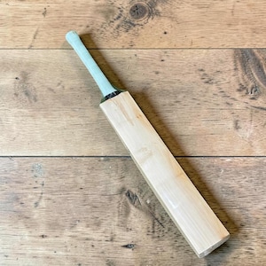 May include: A wooden cricket bat with a light green grip. The bat is made of light-colored wood and is lying on a wooden surface. The bat is a sports equipment item.