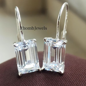 May include: A pair of silver leverback earrings featuring rectangular, emerald-cut clear gemstones. The earrings are presented on a brown surface, with the text "RhombJewels" visible.