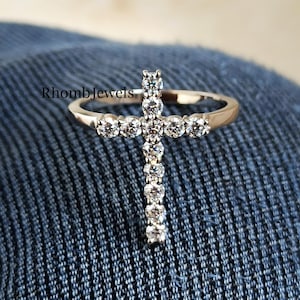 May include: A gold-toned ring featuring a cross design. The cross is adorned with multiple round, clear gemstones. The ring is set against a blue denim background, with the text "RhombJewels" visible.