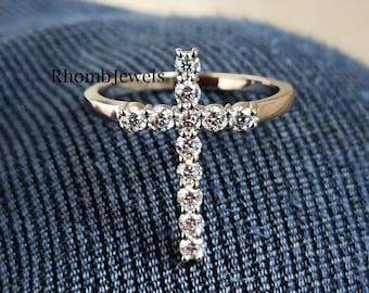 May include: A gold-toned ring featuring a cross design. The cross is adorned with multiple round, clear gemstones. The ring is set against a blue denim background, with the text "RhombJewels" visible.