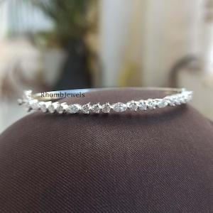 Peut inclure: Un bracelet en argent orné de pierres précieuses claires étincelantes. Le bracelet présente une série de pierres rondes et en forme de marquise, créant un motif continu. Le nom de la marque "RhombJewels" est visible sur le bracelet.