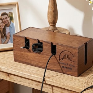 May include: A rectangular dark wood cable management box featuring a laser-engraved design of two birds on a branch with the text "Debbie & Kevin" and "06.09.2026". The box has multiple side slots for routing black charging cables.
