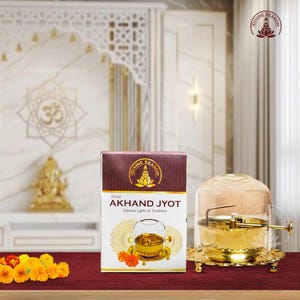 May include: A gold-coloured Akhand Jyot lamp with a glass cover and a decorative base. The product is displayed next to a box with the text "AKHAND JYOT Eternal Light of Tradition". The background features a marble wall and a golden statue.