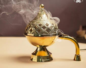 May include: A gold-colored incense burner with a decorative, patterned lid and a curved handle. Wisps of smoke rise from the top, suggesting the burner is in use. The burner sits on a pedestal base, and the overall design is ornate.