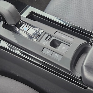 May include: Close-up of a car's center console, featuring a black gear shift lever, buttons for drive modes, and an electronic parking brake. The console has a glossy finish and is surrounded by gray fabric.