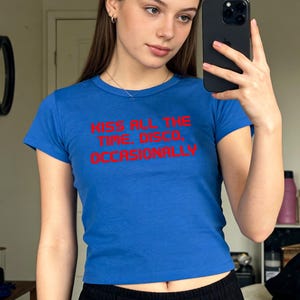 Puede incluir: Una camiseta azul de cuello redondo con el texto "KISS ALL THE TIME. DISCO. OCCASIONALLY" en rojo. La camiseta se combina con pantalones negros. La imagen está tomada frente a un espejo.
