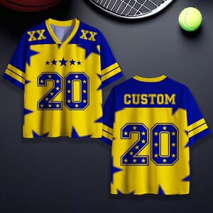 Puede incluir: Dos camisetas deportivas amarillas y azules con el n&uacute;mero 20. La parte delantera de la camiseta tiene el n&uacute;mero 20 en un contorno azul con estrellas. La parte trasera de la camiseta dice "CUSTOM" y tambi&eacute;n tiene el n&uacute;mero 20.