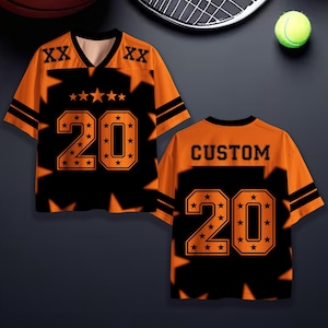 Puede incluir: Camiseta deportiva naranja y negra con el n&uacute;mero 20 y detalles de estrellas. La camiseta tiene cuello en V y mangas cortas. La parte trasera de la camiseta dice "CUSTOM" encima del n&uacute;mero 20.