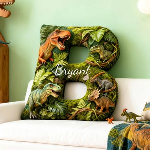 Puede incluir: Un gran cojín con forma de letra "B" cubierto de musgo verde con varias figuras de dinosaurios y el nombre "Bryant" en escritura blanca. El cojín está decorado con helechos y otro follaje, creando un tema prehistórico.